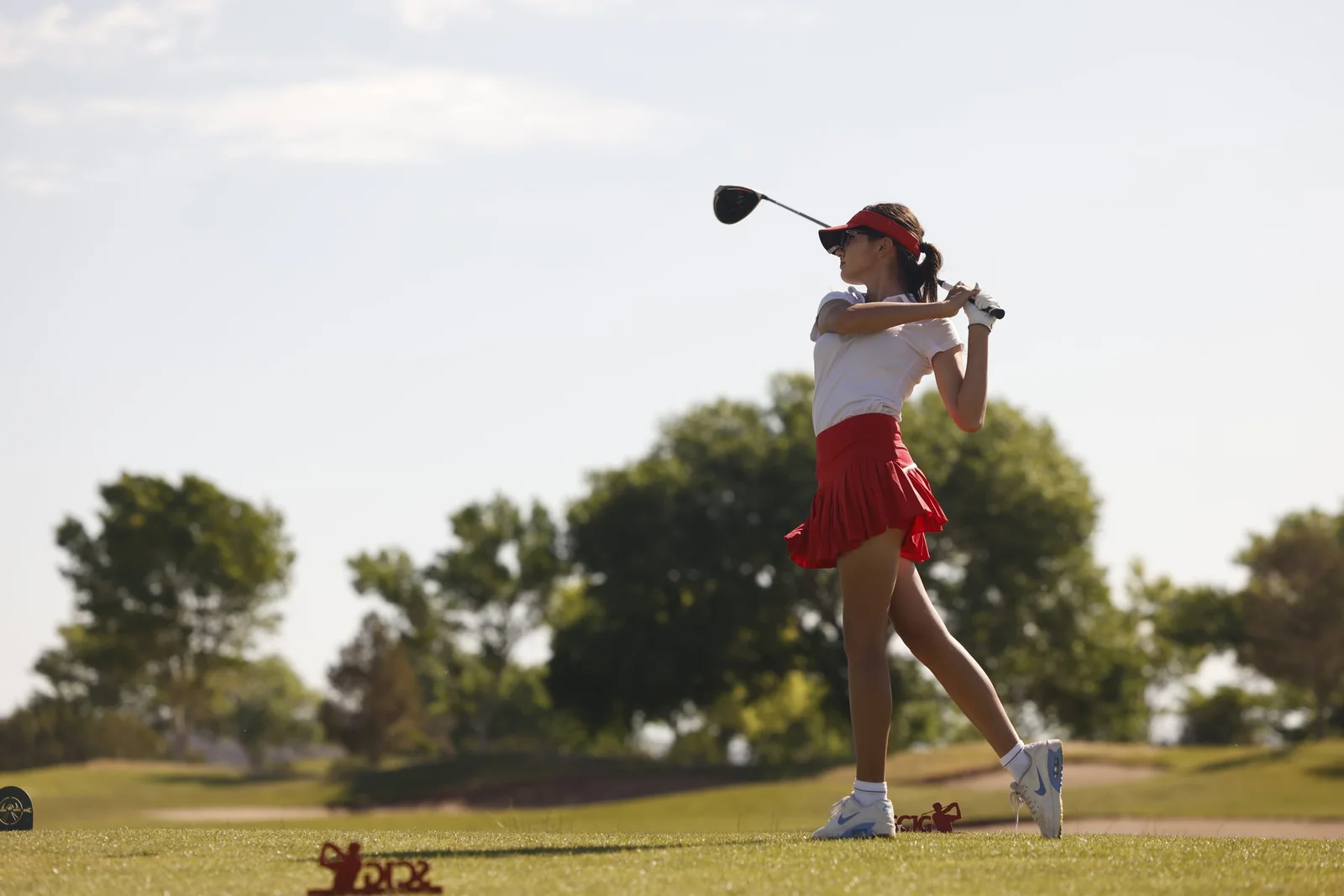 A junior golfer finishes her swing on a tee box under a clear sky.